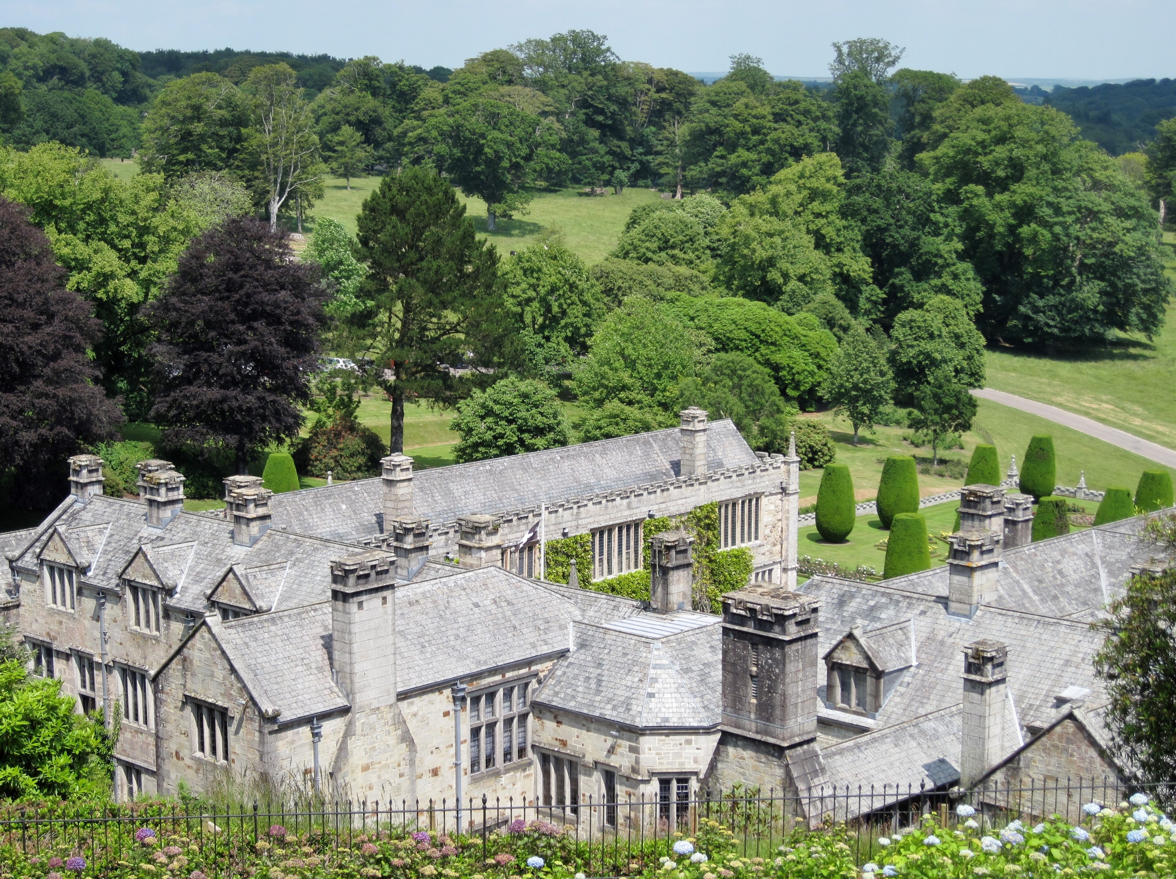 A historic stone building with a complex roof and multiple chimneys, surrounded by lush greenery and a well-maintained lawn.