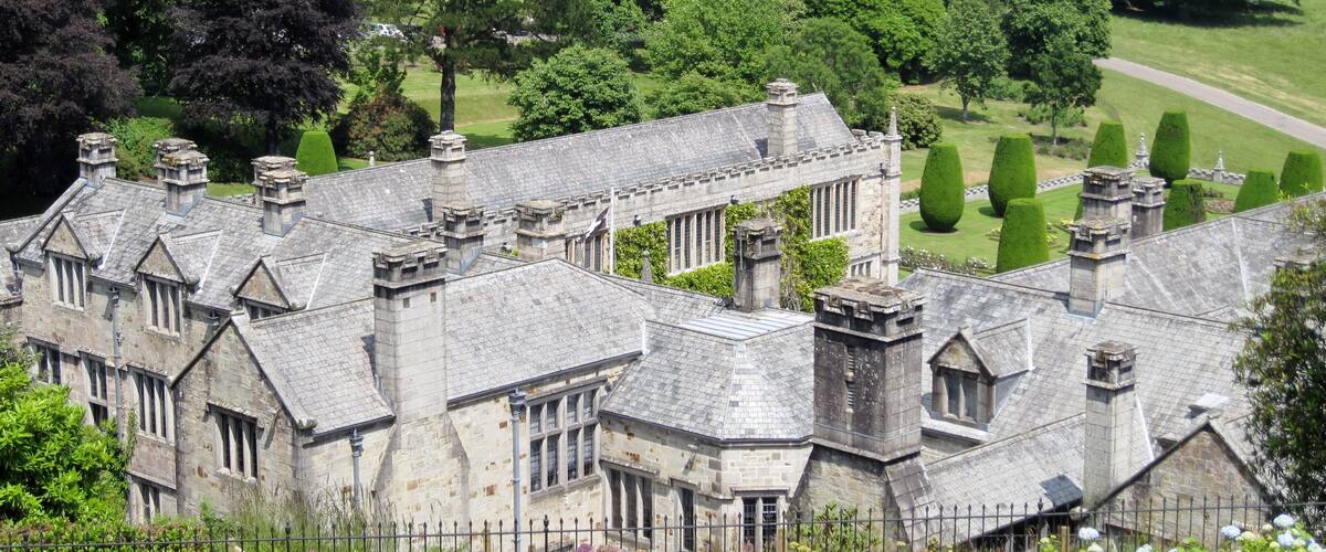 A historic stone building with a complex roof and multiple chimneys, surrounded by lush greenery and a well-maintained lawn.