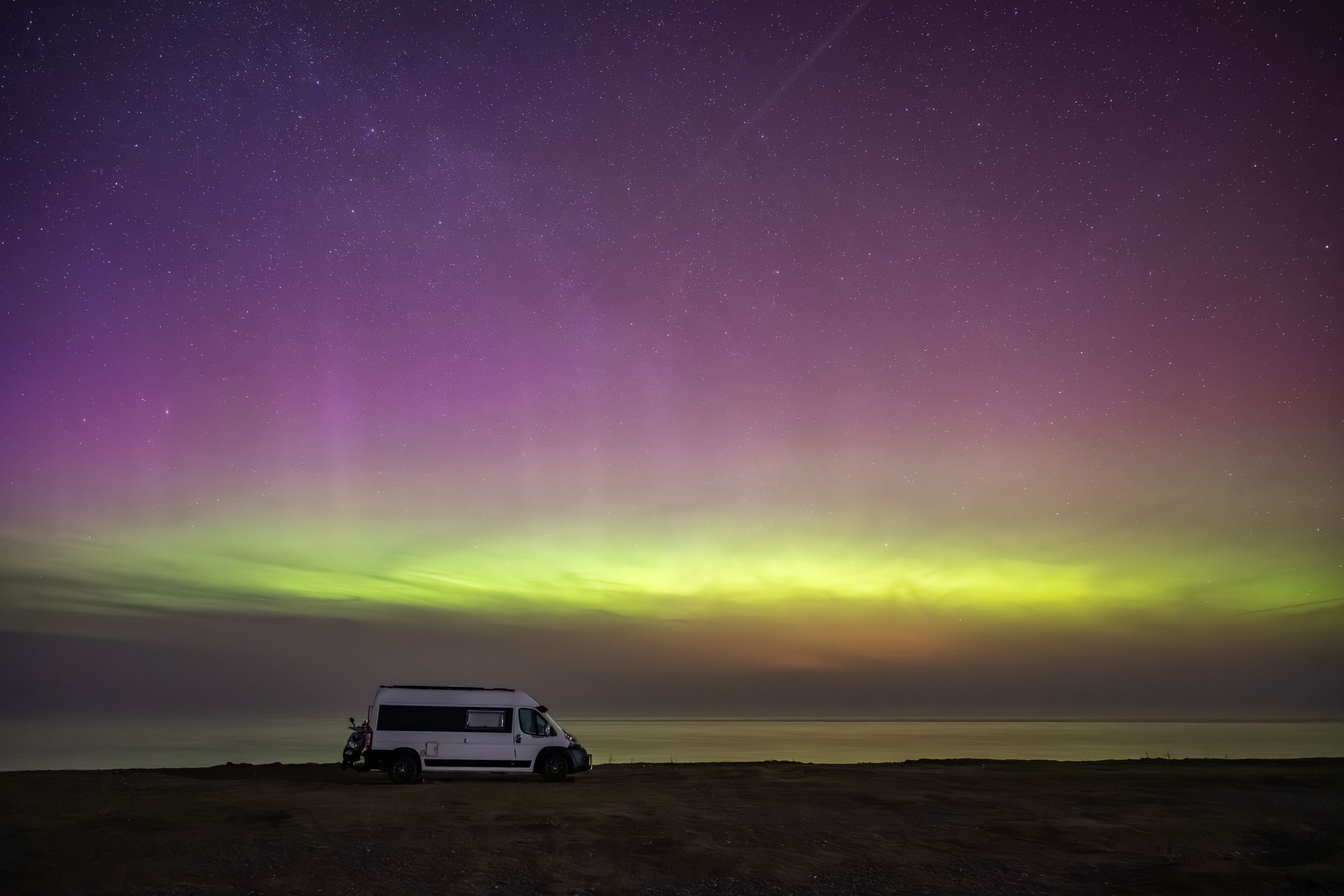 A white van parked on a beach under a starry night sky with the aurora borealis.