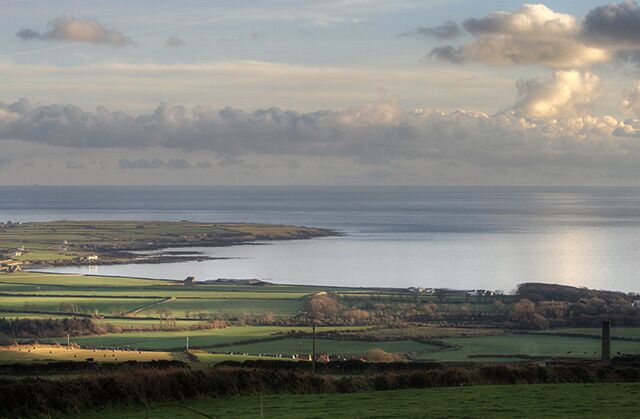 A coastal landscape with fields, a body of water, and distant hills.