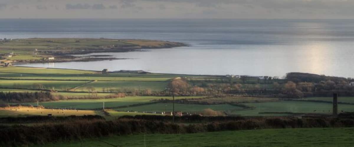 A coastal landscape with fields, a body of water, and distant hills.