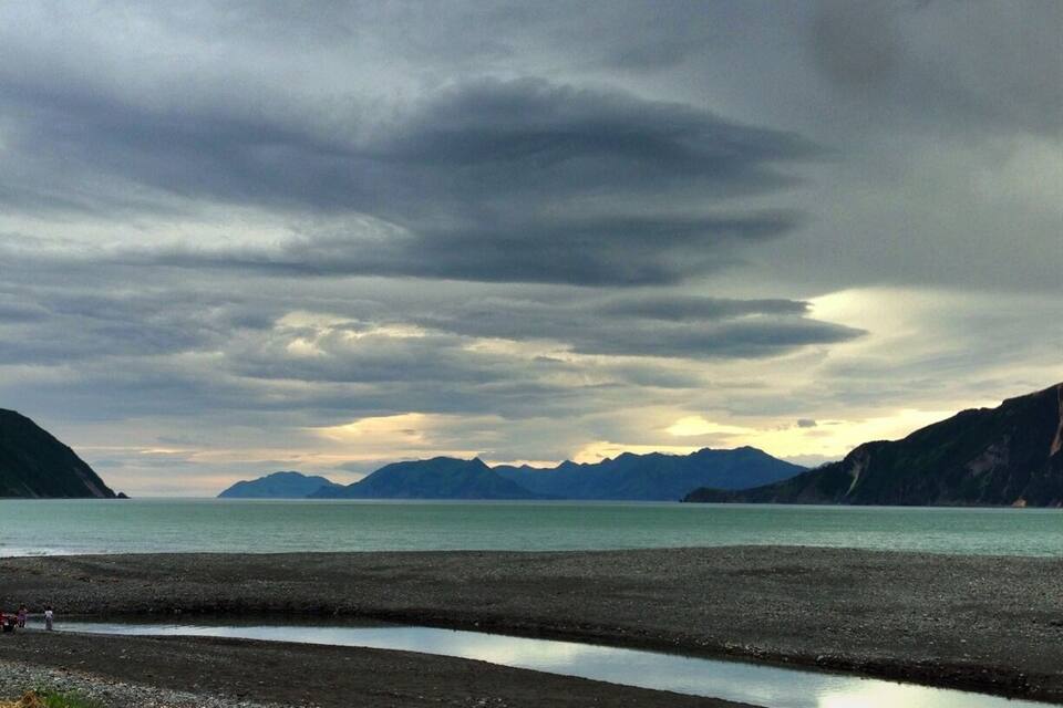 A coastal landscape with a rocky beach, calm sea, and distant mountains under a cloudy sky.