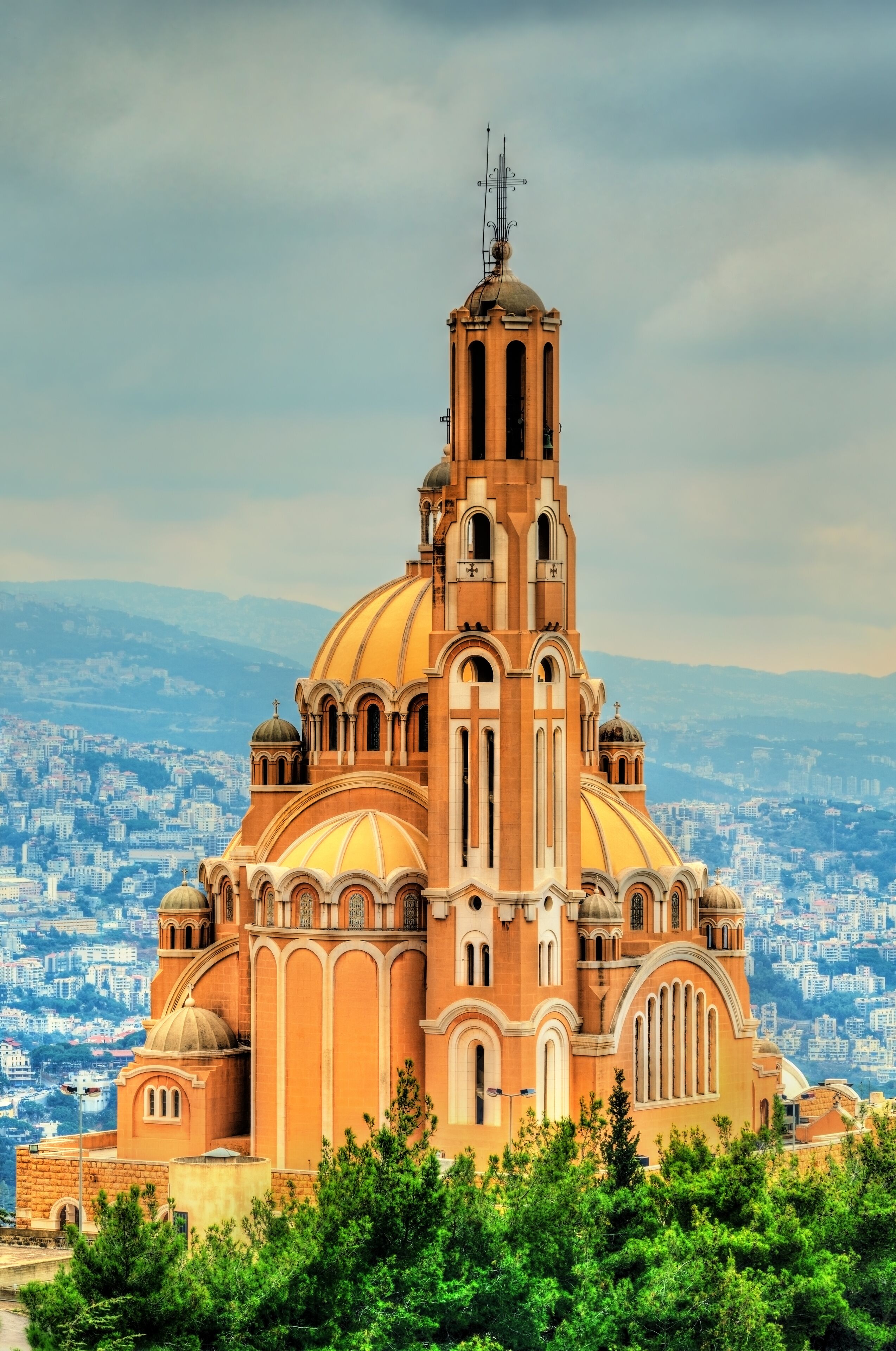 A large church with a tall bell tower and multiple domes, surrounded by greenery and a cityscape in the background.