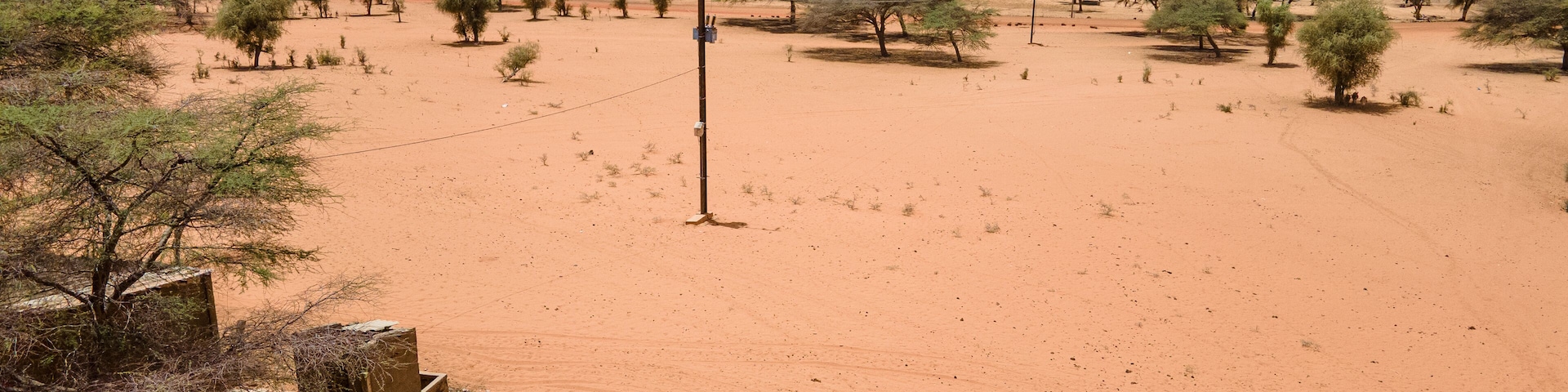 Aerial view of arid lands punctuated by sporadic trees, a simple structure, and individuals on donkey-drawn carts under a vast sky, Khol Khol, Louga Region, Senegal.