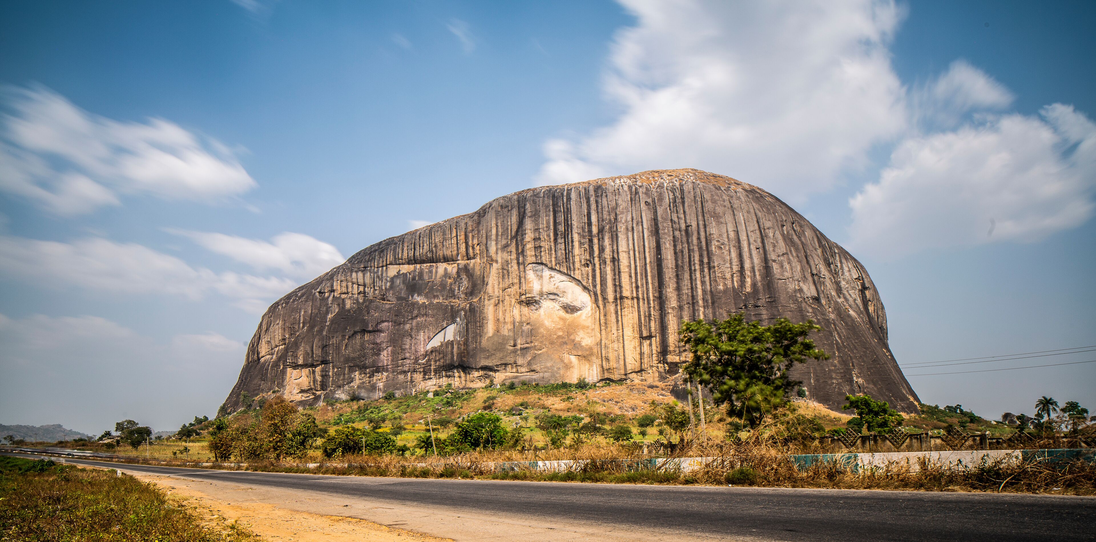 A large rock formation with a carved face, surrounded by greenery and a road.