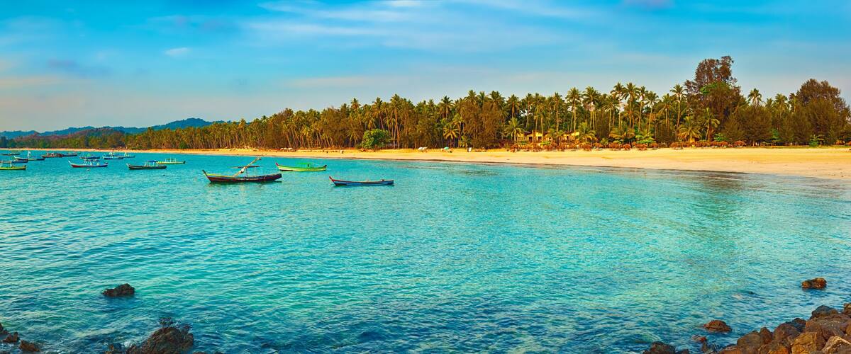 A beach with clear turquoise water, small boats, and a line of palm trees.