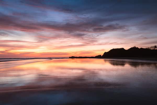 Una playa al atardecer, con un mar tranquilo que refleja el cielo.