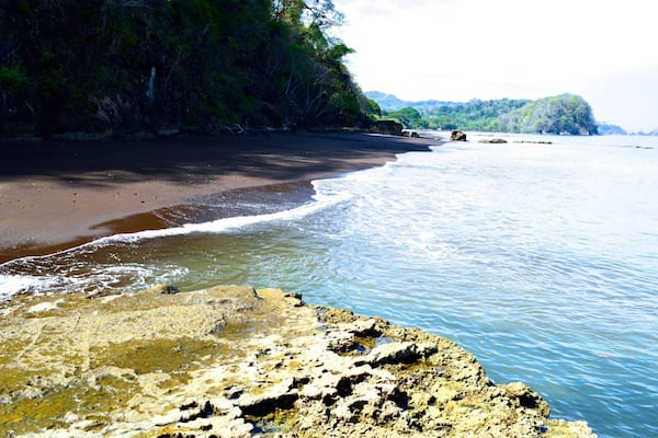 Une plage avec un rivage rocheux au premier plan, une étendue de sable et une colline boisée en arrière-plan.