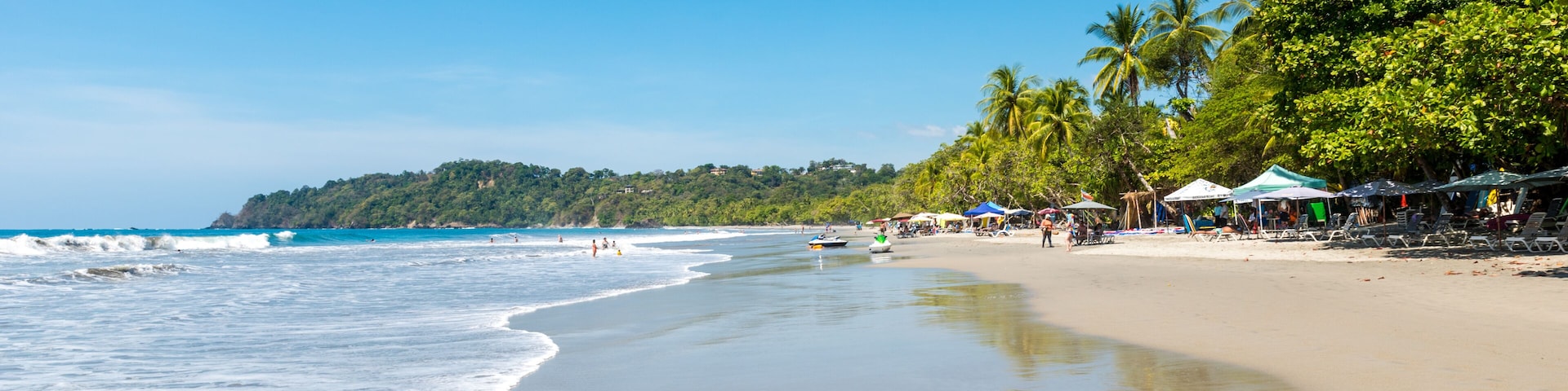 Uma praia com águas azul-claras, areal arenoso, palmeiras e guarda-sóis.