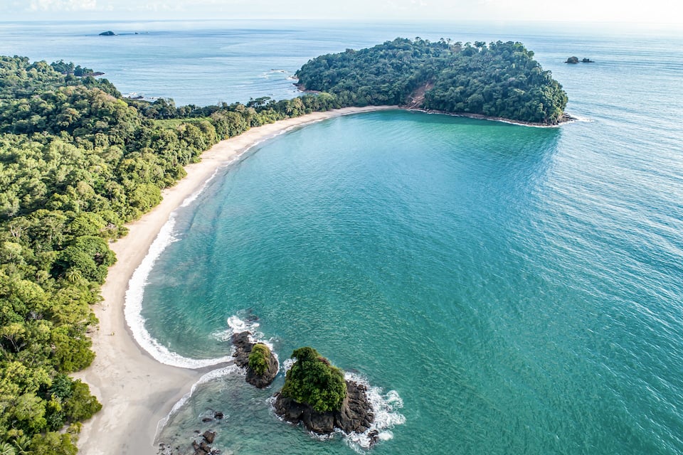 A coastal landscape with a sandy beach, clear turquoise waters, and a lush green island.