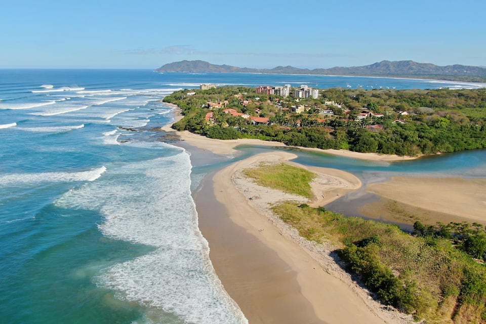 Paisagem costeira com praia arenosa, um rio sinuoso e área residencial com edifícios numa encosta.