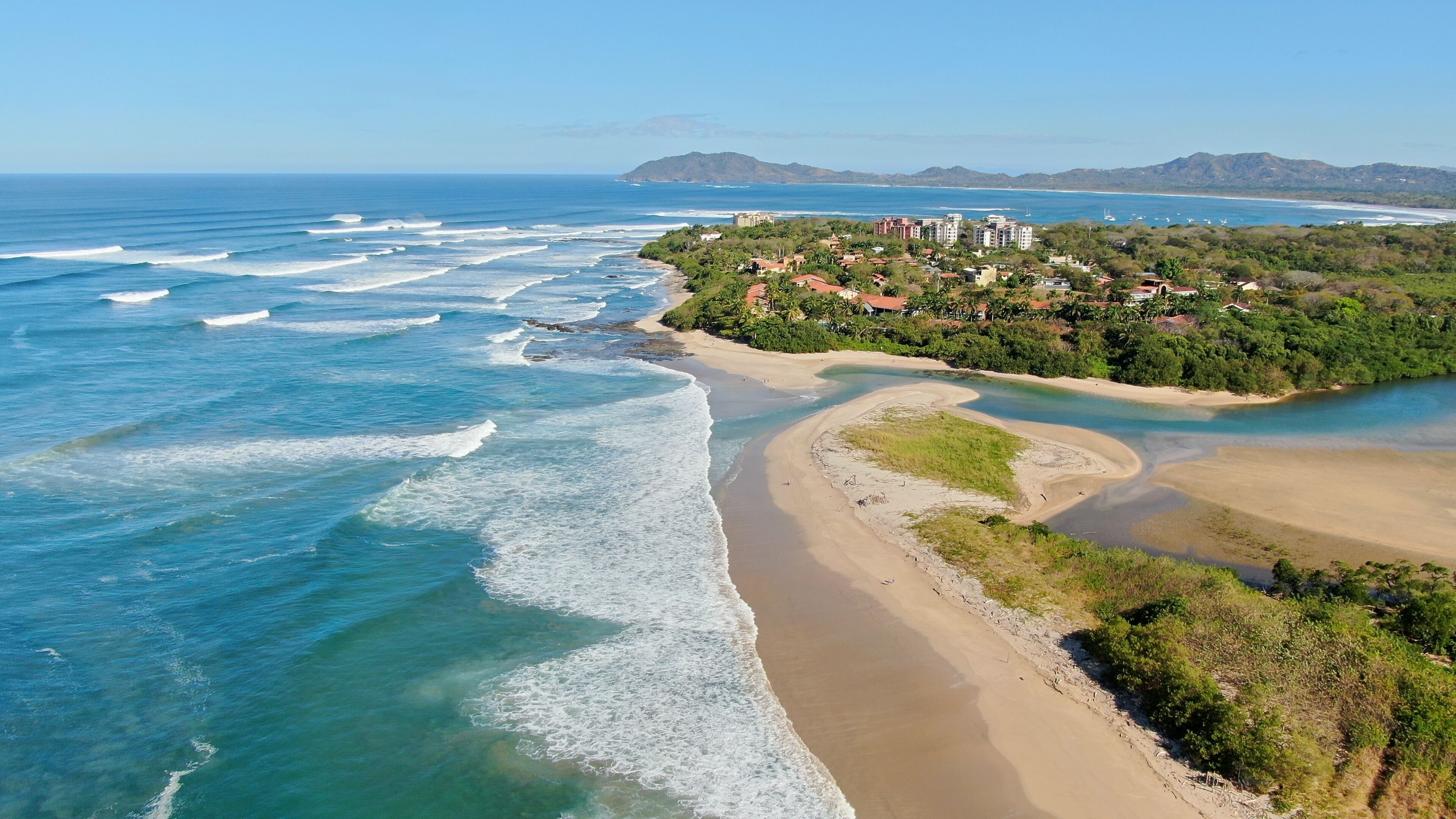 A coastal landscape with a sandy beach, a winding river, and a residential area with buildings on a hillside.