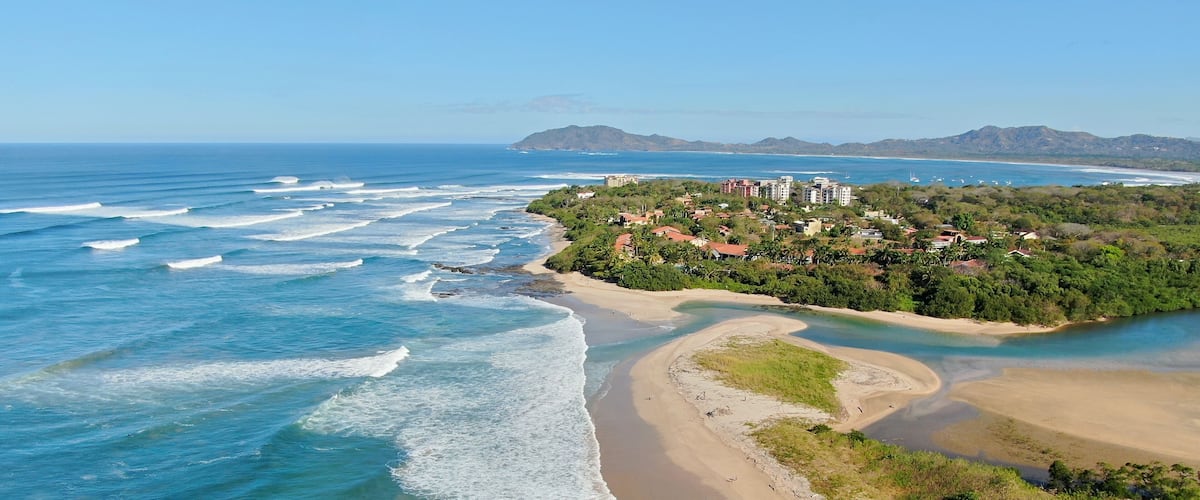 A coastal landscape with a sandy beach, a winding river, and a residential area with buildings on a hillside.