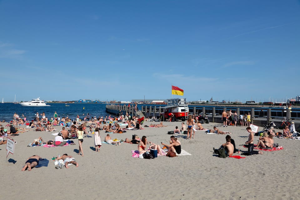 Ein überfüllter Strand mit badenden und sonnenden Menschen, ein Steg mit Booten und eine wehende Flagge.