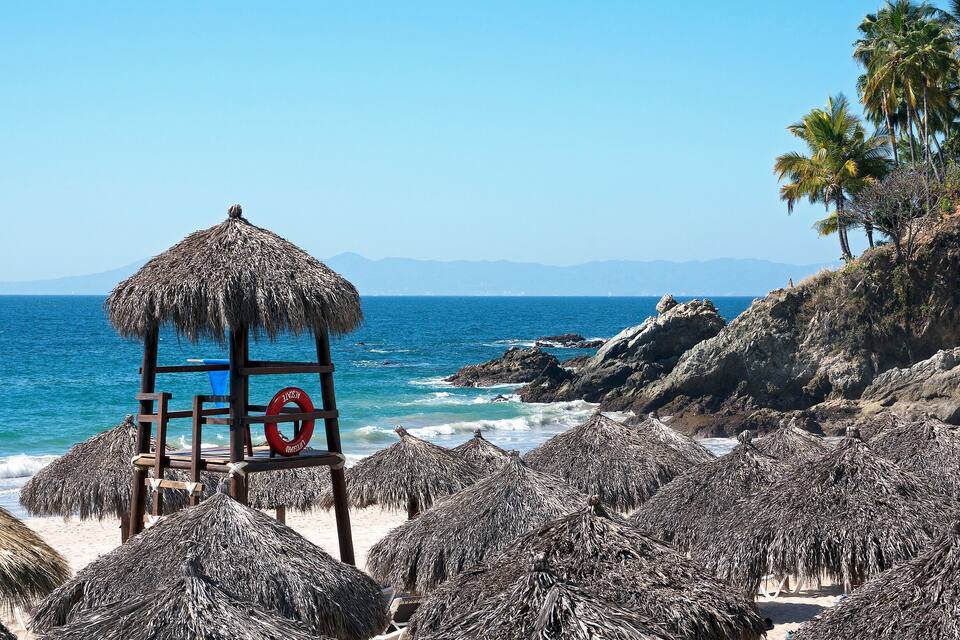 Uma praia com guarda-sóis de palha, boia salva-vidas, costa rochosa e palmeiras.
