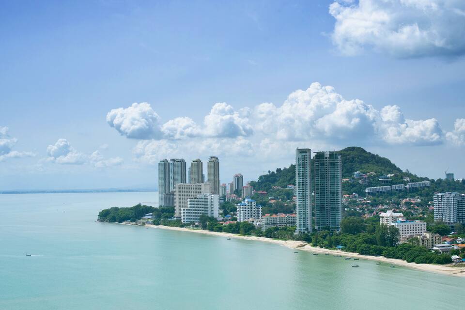 Paisagem costeira com edifícios altos, praia e uma colina com vegetação.
