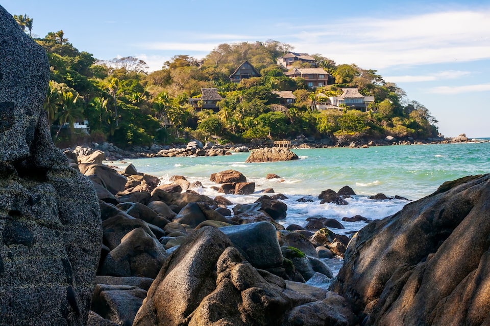 A rocky shoreline with large boulders, a clear blue sea, and a hilltop with houses and lush greenery.