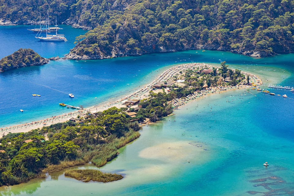 An aerial view of a coastal area with clear turquoise waters, a sandy beach, and lush green hills.