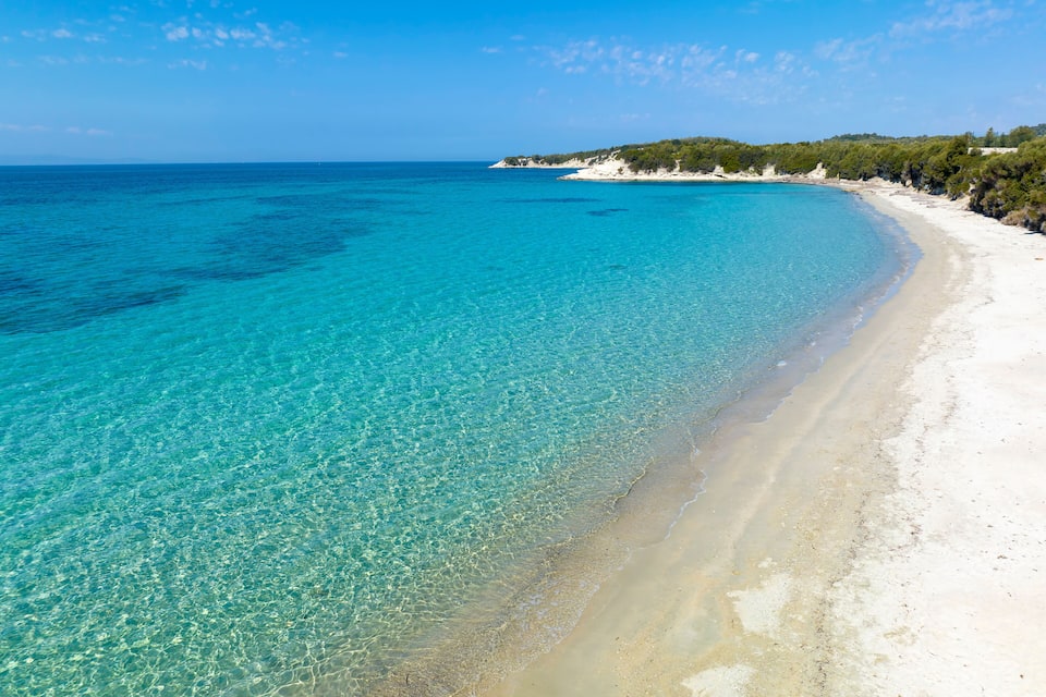 A clear turquoise sea, white sandy beach, and a forested hill in the distance.