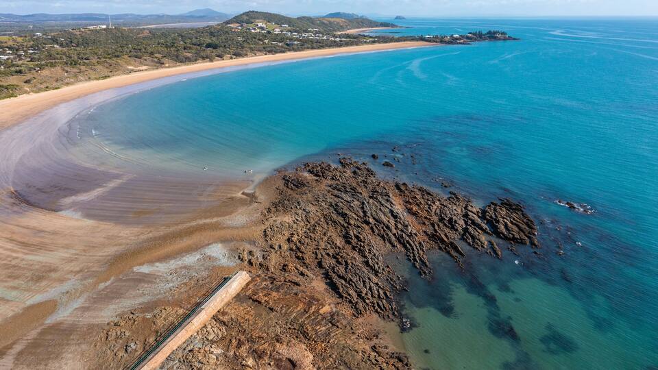 An aerial view of a coastal area with a sandy beach, clear blue waters, and rocky outcrops.