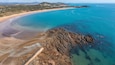 Luchtbeeld van een kustgebied met een zandstrand, helderblauw water en rotsformaties.