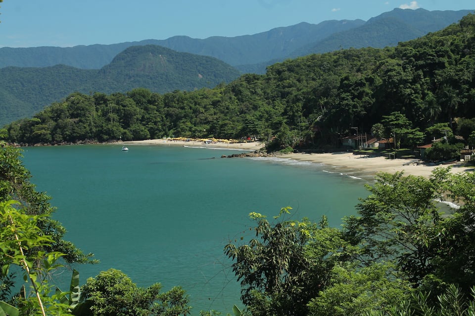 Paisagem costeira com uma praia, águas turquesa cristalinas e colinas verdes exuberantes ao fundo.