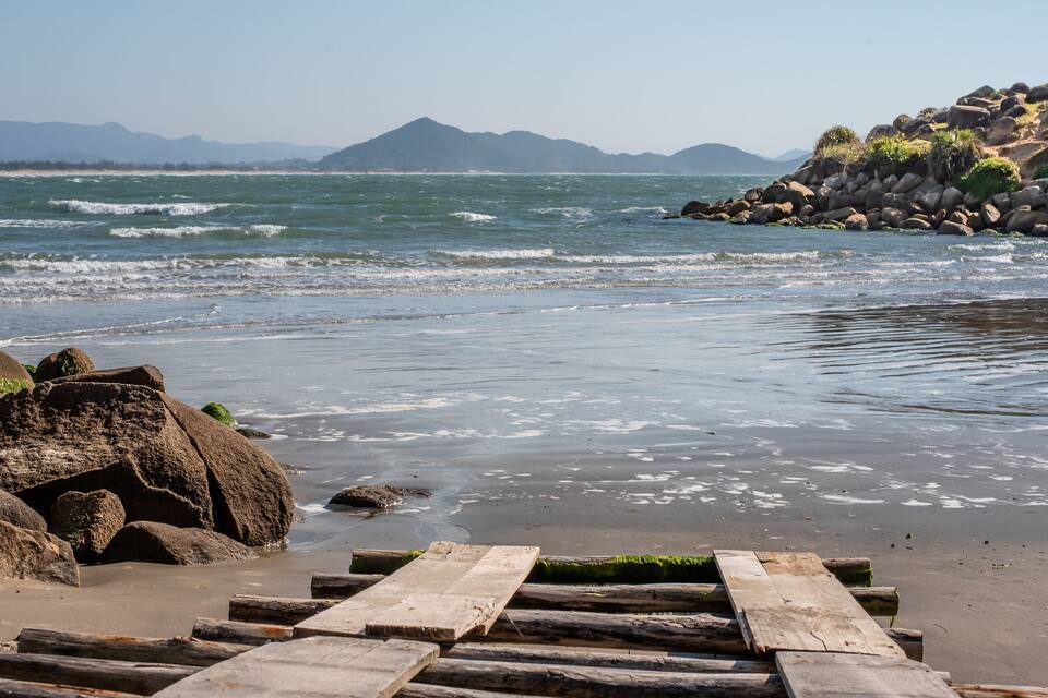 Una playa con afloramientos rocosos, pasarelas de madera y montañas al fondo.