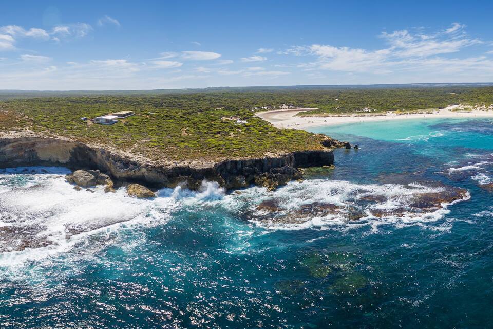 Un paisaje costero con un prominente afloramiento rocoso rodeado de aguas turquesas, una playa de arena y una colina verde distante.