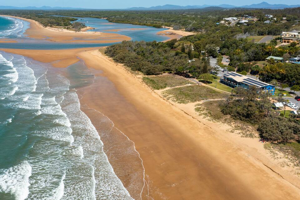 Un paysage côtier avec une plage de sable, une rivière et une zone résidentielle comprenant des maisons et un bâtiment doté d’un toit bleu.