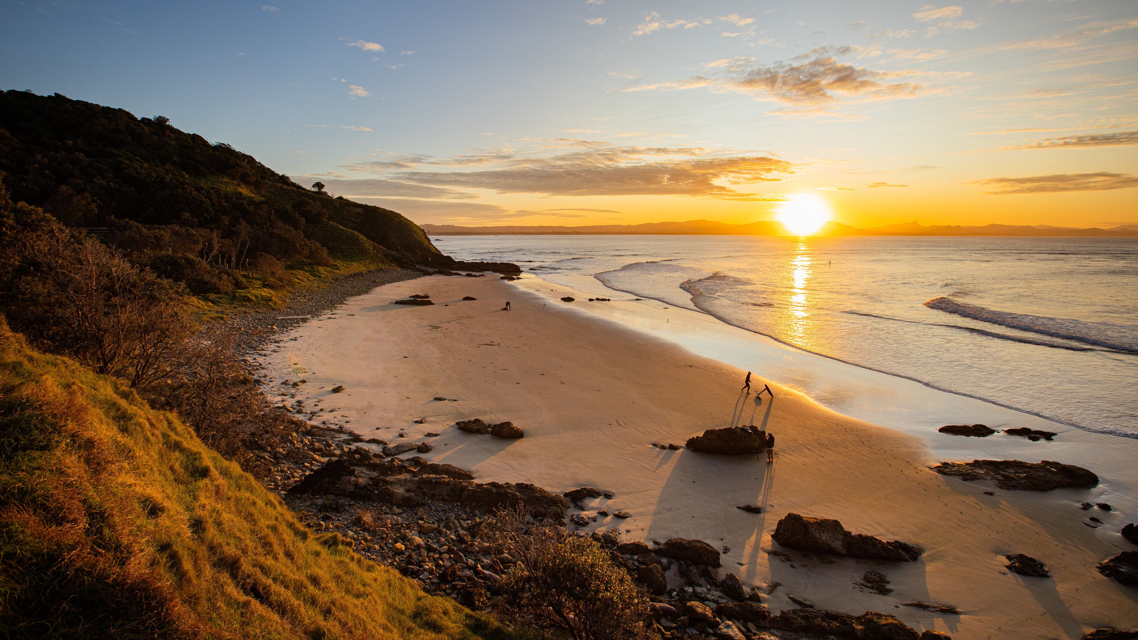 A beach with a sunset, rocky shoreline, and grassy hills.