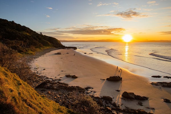 A beach with a sunset, rocky shoreline, and grassy hills.