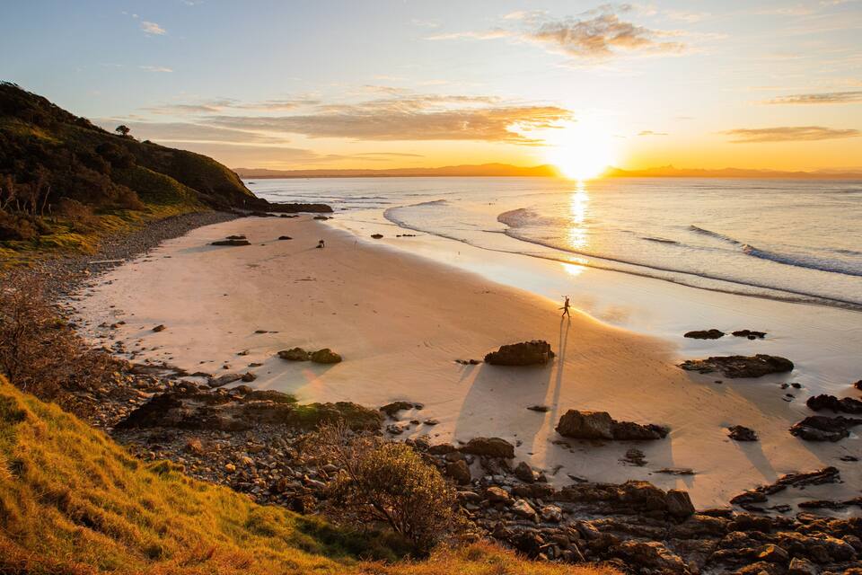 A beach with a sunset, rocky shoreline, and grassy hills.