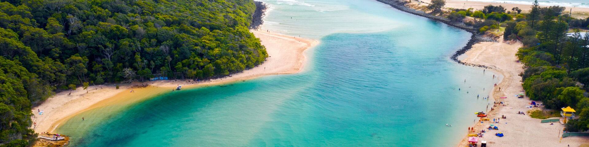 A coastal landscape with a clear turquoise bay, sandy beach, and dense forested hills.