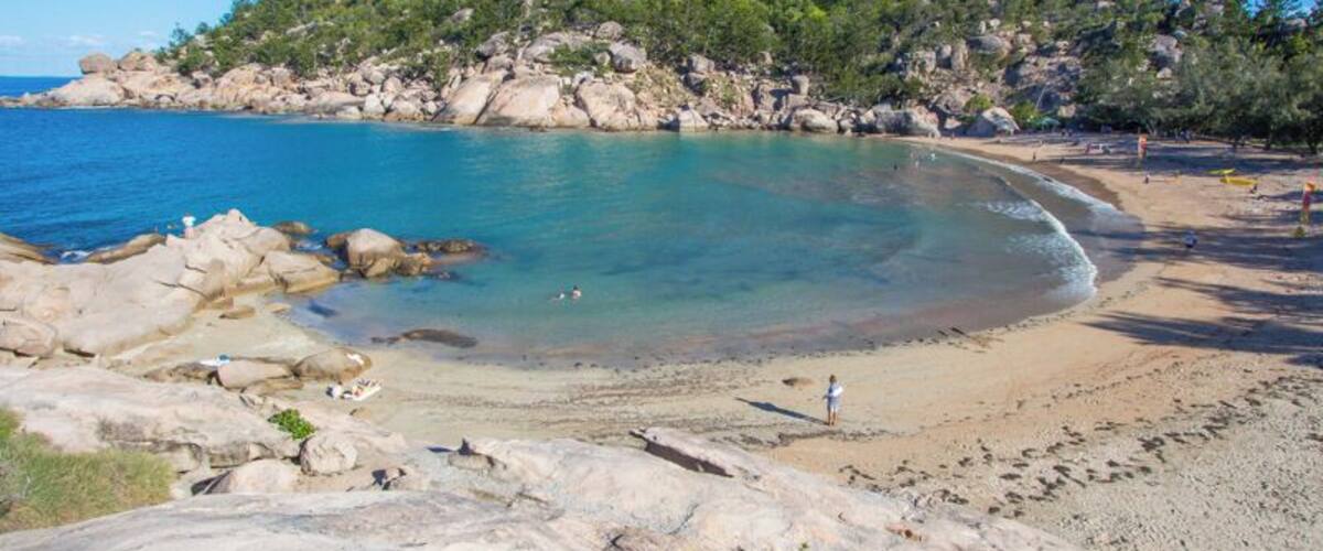A beach with clear turquoise water, rocky shoreline, and a forested hill in the background.