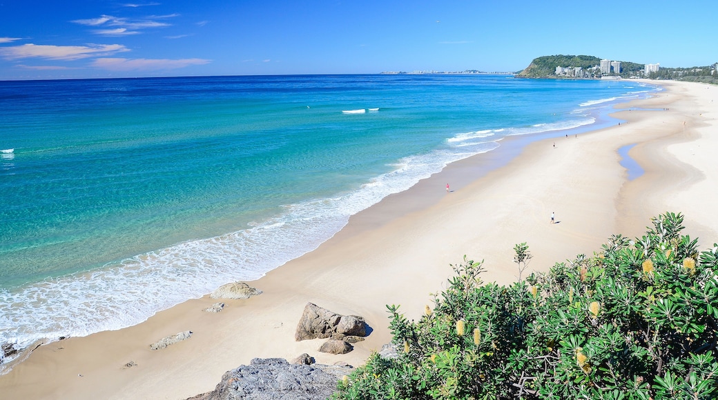 A beach with clear turquoise waters, a sandy shore, and a distant hill with buildings.