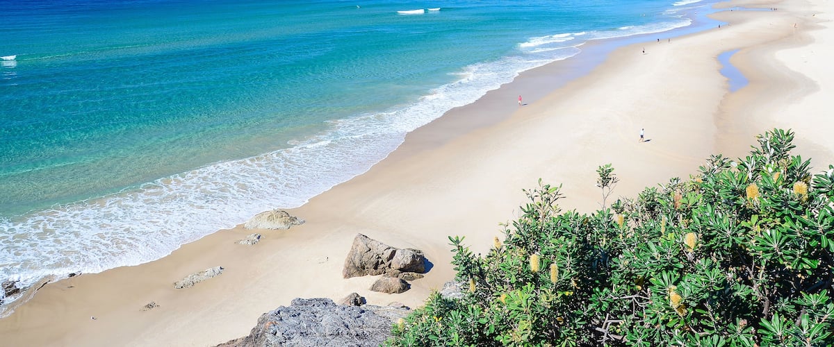 A beach with clear turquoise waters, a sandy shore, and a distant hill with buildings.