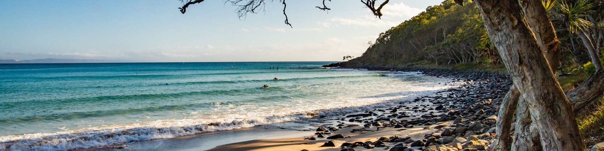 A rocky beach with clear turquoise water, a tree-lined shoreline, and distant mountains.