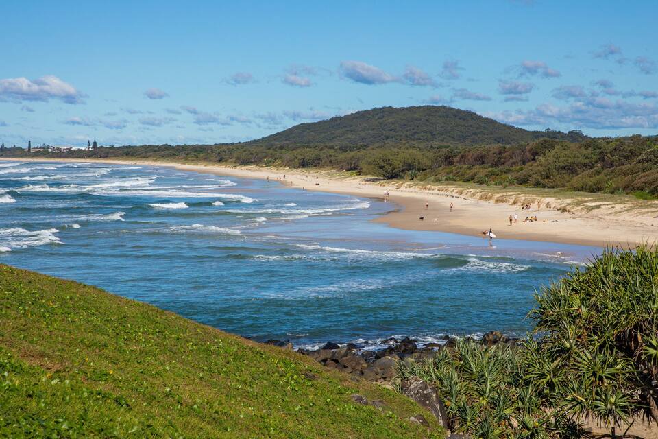 Une plage avec des vagues, une rive sablonneuse et une colline verdoyante en arrière-plan.