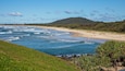 Uma praia com ondas, uma faixa de areia e uma colina verde ao fundo.