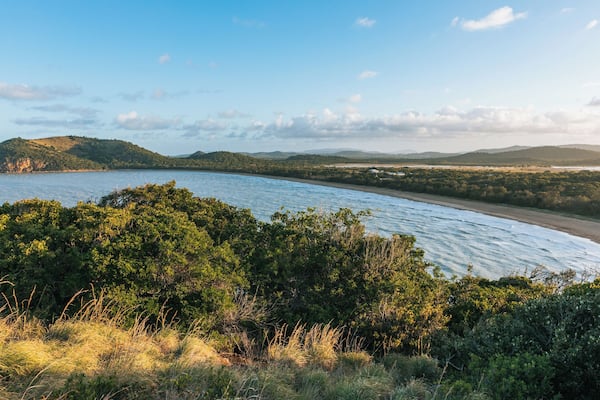 A coastal landscape with a beach, dense greenery, and distant hills.