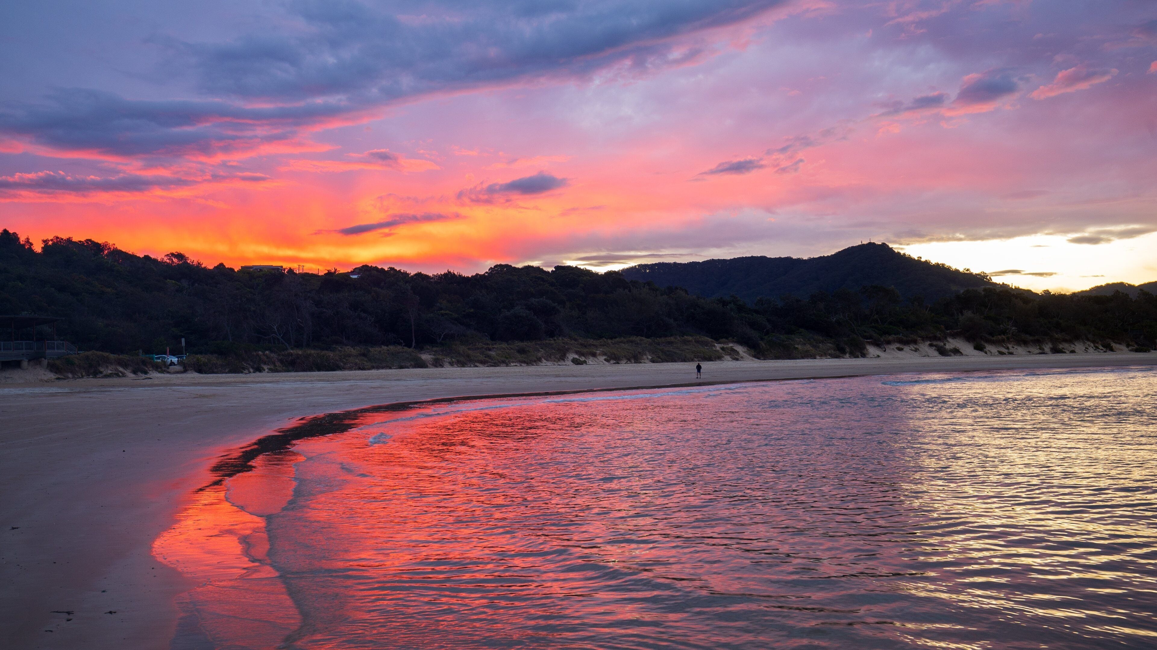 A beach with a sunset, calm water reflecting the sky, and a forested hill in the background.