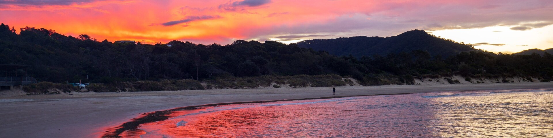 A beach with a sunset, calm water reflecting the sky, and a forested hill in the background.