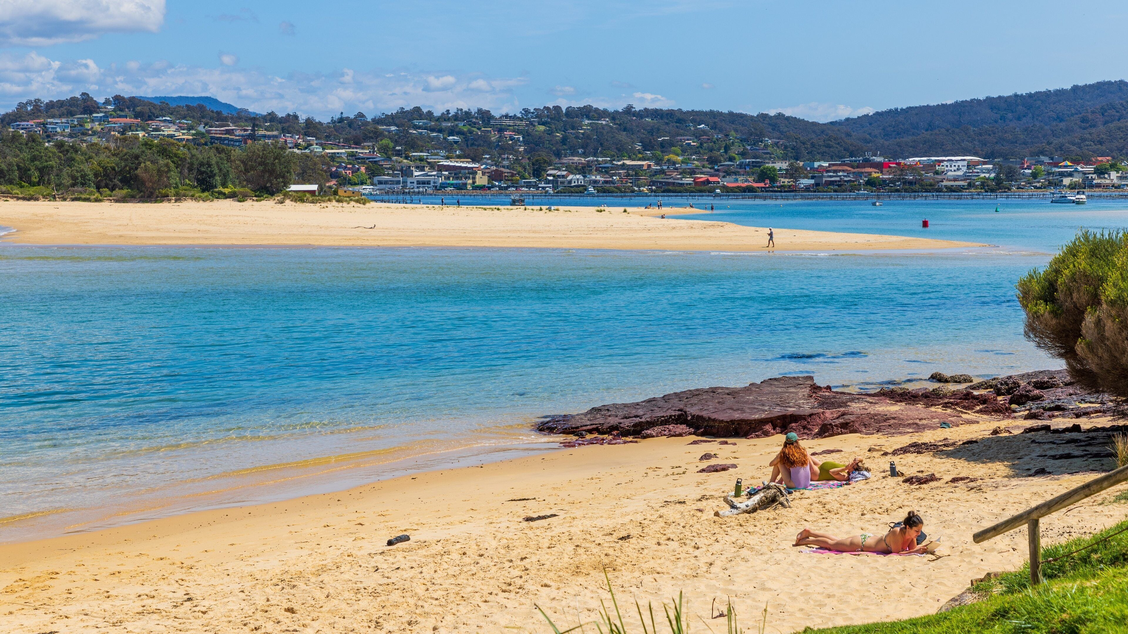 A sandy beach with clear blue water, a rocky outcrop, and a town in the background.