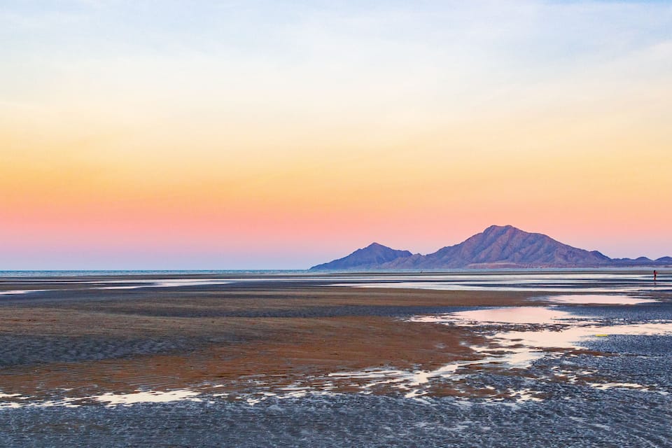 Un paisaje costero con una montaña al fondo y una persona de pie en la playa.