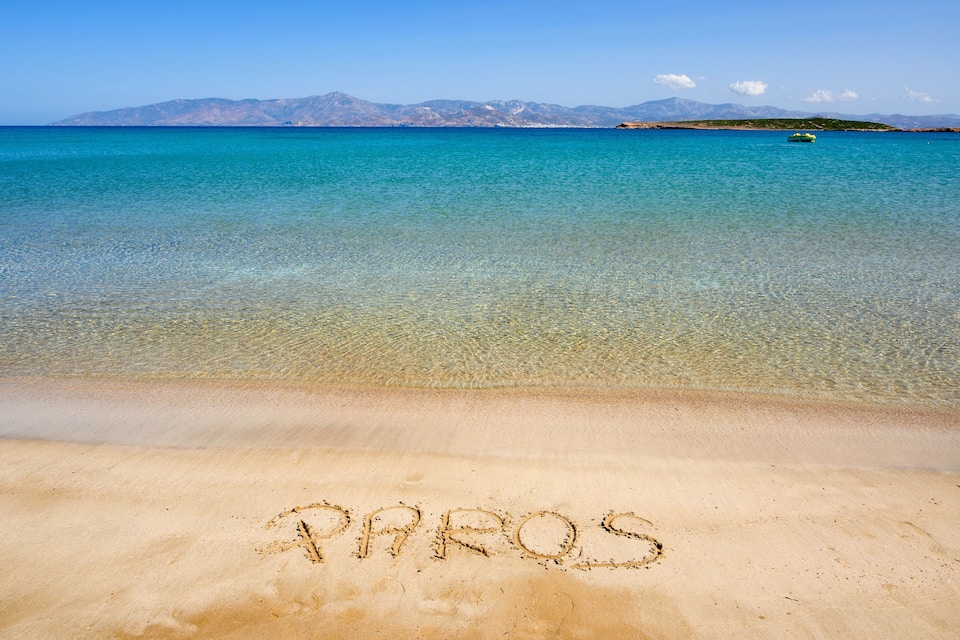 Ein Strand mit 'PAROS' im Sand geschrieben und einem klaren blauen Meer mit Bergen im Hintergrund.