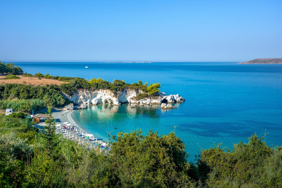 A coastal landscape with a clear blue sea, rocky cliffs, and a beach with umbrellas.