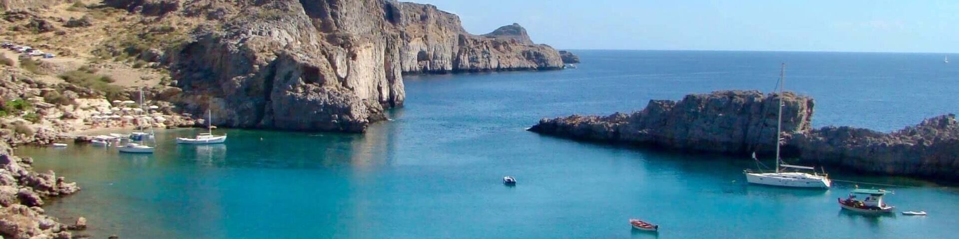 A coastal landscape with rocky cliffs, clear turquoise waters, and several boats anchored near the shore.