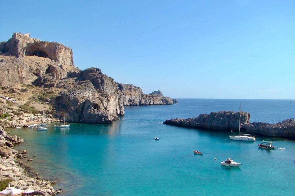 A coastal landscape with rocky cliffs, clear turquoise waters, and several boats anchored near the shore.