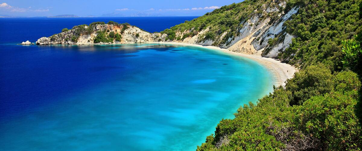 A coastal landscape with a clear blue sea, a sandy beach, and a green hillside.