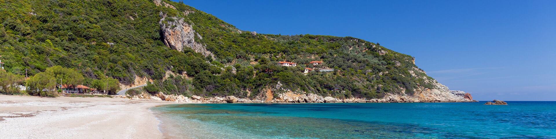 A clear beach with turquoise water, a sandy shore, and a green hillside with buildings.